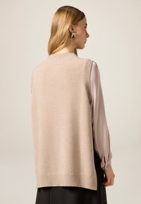 Beiger ärmelloser Strickpullover mit geripptem Ausschnitt und Saum, darunter eine hellgraue Bluse mit Puffärmeln getragen. Goldene geometrische Ohrringe.