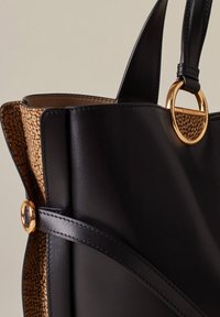 Schwarze Leder-Umhängetasche mit einem strukturierten beigen Innenfutter, verziert mit einem goldenen runden Hardware-Detail und dekorativen Lederstrapselementen.