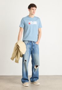 Šviesiai mėlynas medvilninis marškinėliai su baltu ir raudonu "CALVIN KLEIN" logotipu, derinami su nusidėvėjusiais šviesiai mėlynais džinsais ir smėlio spalvos bombone. 