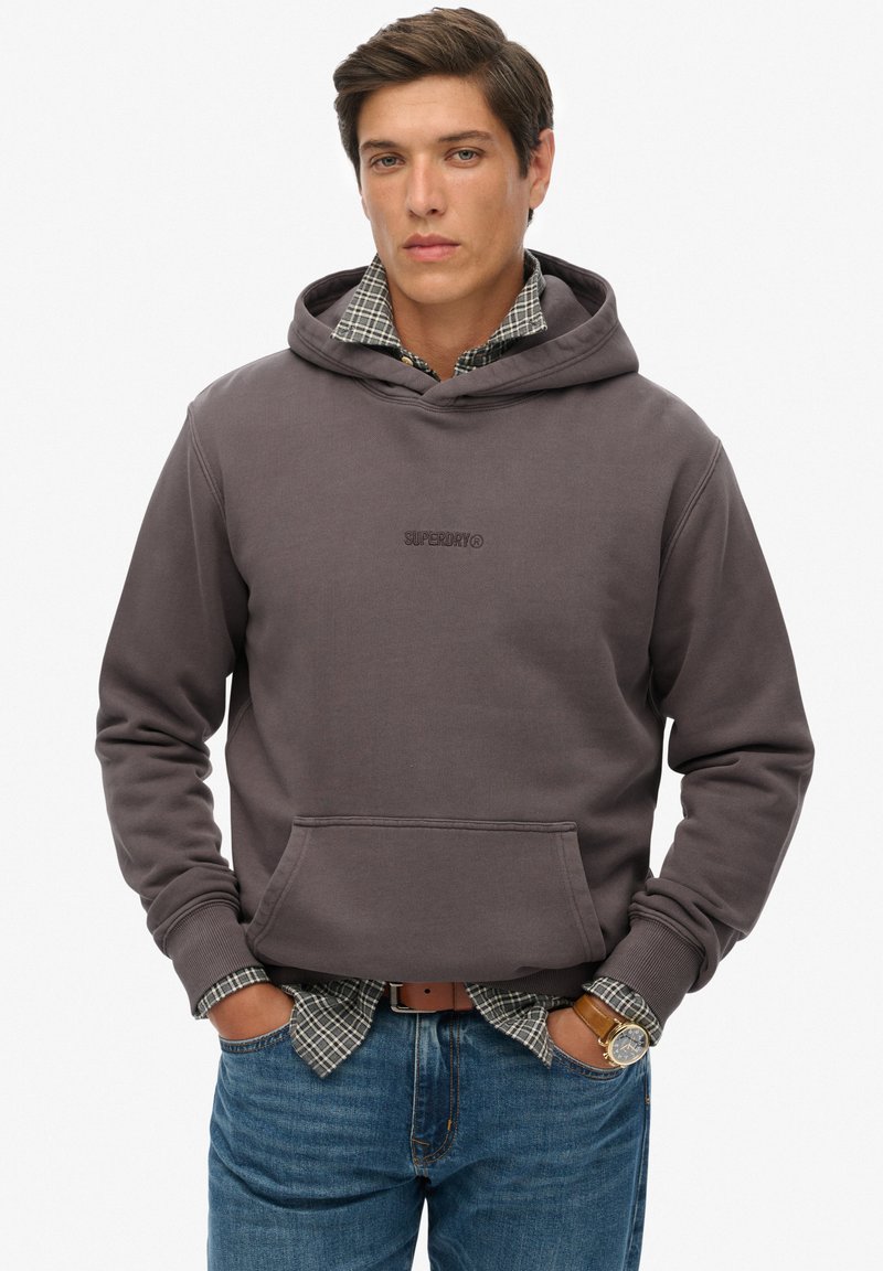 Sudadera con capucha de color gris oscuro, con bolsillo frontal y logotipo bordado. Lleva encima una camisa de cuadros, combinada con jeans azules. Diseño casual.