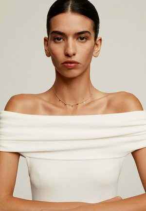 Top blanco de canalé sin hombros, textura suave, diseño ajustado, mostrado en una modelo con un collar dorado sencillo, fondo neutro.