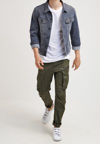 Džínová bunda v tmavě šedé, bílý tričko, olivově zelené cargo kalhoty s kapsami a bílé tenisky, představující neformální outfit.