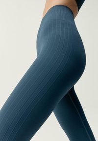 Aukštos juosmens sportinės kelnės tamsiai žalsva spalva, su šonkaulinio rašto tekstūra ir vertikaliomis linijomis, turinčios kontrastingus siūlus šonuose.