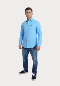 Camisa de botón azul claro con mangas largas y un logo bordado en el pecho, combinada con vaqueros oscuros y zapatillas.