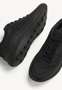 Paire de chaussures de sport noires avec semelles texturées, dessus en tissu tricoté et design à lacets sur fond blanc.