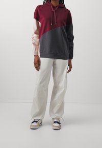 Sudadera a bloques de color con paneles en burdeos, rosa claro y gris oscuro; cuenta con capucha con cordones y puños acanalados. Combinada con pantalones blancos.