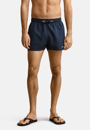 Man met blote torso die marineblauwe zwembroek en bruine teenslippers draagt, staand tegen een eenvoudige lichte achtergrond.