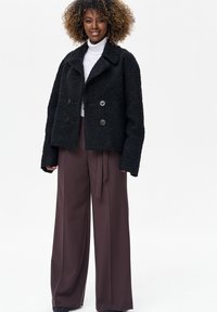 Czarna teksturowana kurtka z podwójnymi guzikami, zestawiona z brązowymi, szerokimi spodniami z wysokim stanem oraz białym golfem pod spodem.