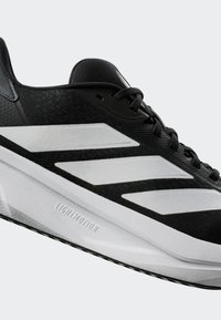 Black athletic shoe with white stripes, textured upper, lightweight midsole, and rubber outsole. Features a lace-up design and branding.