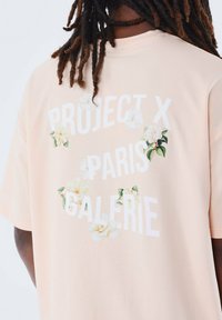Lichtroze katoenen t-shirt met een bloemig ontwerp en de tekst: "PROJECT X PARIS GALERIE" in het wit. Korte mouwen, relaxte pasvorm.