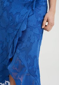 Modré šaty s květinovou výšivkou, průsvitnou vrstvou a asymetrickým lemem. Textura je měkká s vyvýšenými detaily. Ruka držící látku.