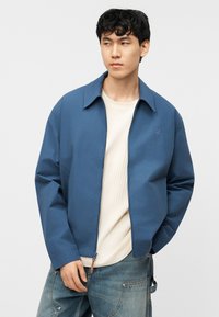 Νέος άνδρας που φοράει μπλε μπουφάν με φερμουάρ πάνω από λευκή ριγωτή μπλούζα και άνετο μπλε τζιν, στέκεται με το ένα χέρι στην τσέπη.