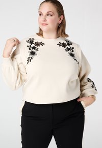 Beige sweatshirt met zwarte bloemrijke borduursels op de schouders en mouwen, ontspannen pasvorm, lange opgepompte mouwen. Gecombineerd met zwarte broek.