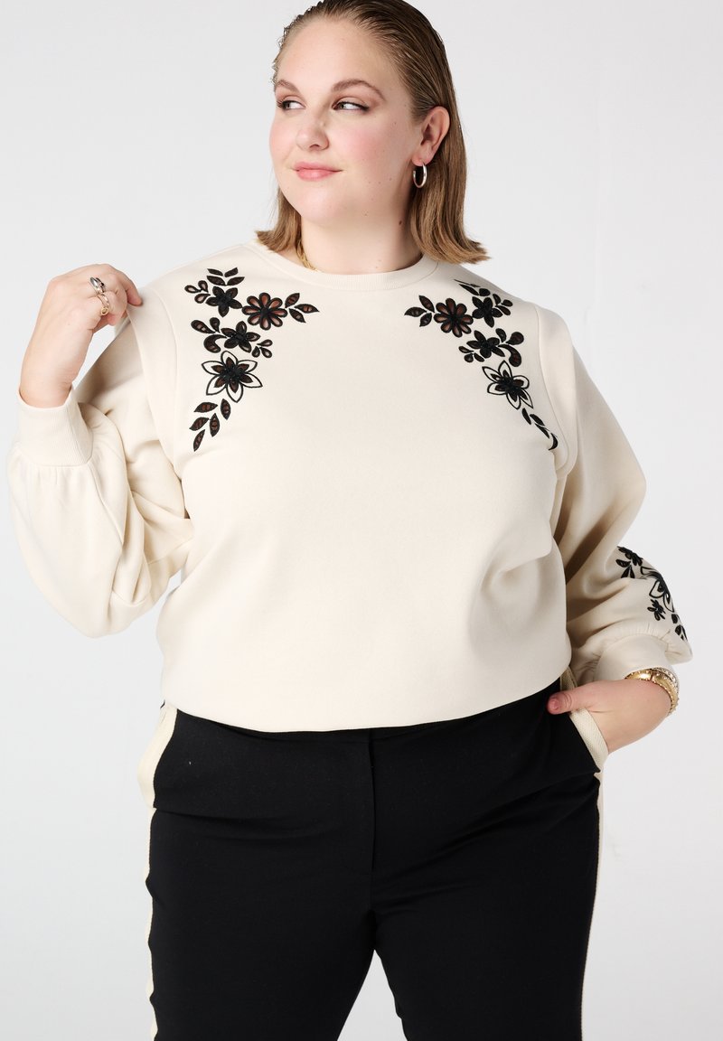 Beige sweatshirt met zwarte bloemrijke borduursels op de schouders en mouwen, ontspannen pasvorm, lange opgepompte mouwen. Gecombineerd met zwarte broek.