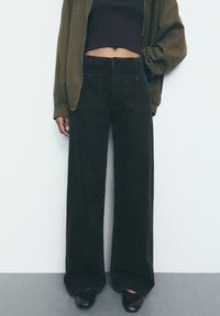 Schwarze weite Hose aus strapazierfähigem Denim-Stoff mit Fronttaschen und hoher Taille, kombiniert mit einer olivgrünen Zip-Jacke.