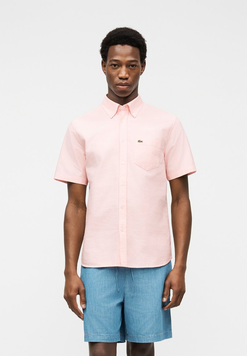 Homme portant une chemise à manches courtes rose pâle avec une poche poitrine et un short en denim bleu, debout devant un fond clair uni.