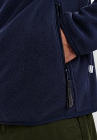 Chaqueta de forro polar azul marino con tela texturizada, bolsillo frontal con cremallera y tirador de cremallera de marca. Presenta un dobladillo ajustado y puños en las muñecas.