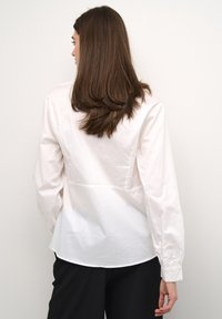 Camicia bianca con bottoni e maniche lunghe, realizzata in cotone liscio, con un design aderente sul retro. Presenta un orlo curvo e polsini con bottone.
