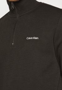 Pull noir à manches longues avec zip au quart et logo blanc "Calvin Klein" sur la poitrine gauche.