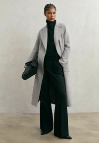 Dolga siva volnena bunda preko črnega majice z visokim ovratnikom in širokih črnih hlač, združena s črno torbico. Gladek tekstura in krojen dizajn.