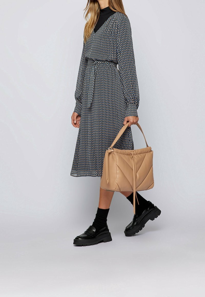 Femme portant une robe midi à manches longues à motifs, des chaussettes noires et des mocassins, portant un grand sac à main beige dans sa main gauche.