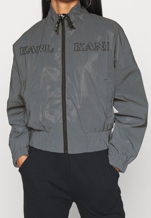 Persoon draagt een grijze jas met ritssluiting met "KARL KANI" in het zwart geborduurd op de borst, handen in de zakken van een zwarte broek.