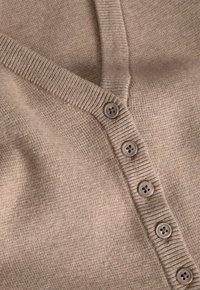 Close-up van beige gebreide cardiganstof met geribbelde randen en vijf bijpassende ronde knoppen die verticaal zijn uitgelijnd.