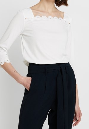 Vrouw draagt een witte blouse met een vierkante halslijn met geschulpte rand, driekwart mouwen en een zwarte broek met hoge taille en ceintuur, hand in de zak.