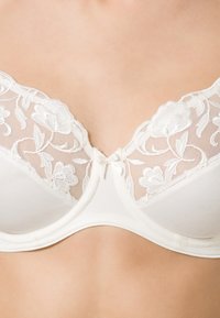 Reggiseno bianco con un pannello in pizzo floreale trasparente nella parte superiore, coppe lisce e un piccolo fiocco al centro. Tessuto morbido e design strutturato.