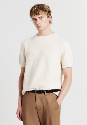 Maglione a maniche corte in crema con texture a maglia, con scollo rotondo, abbinato a pantaloni beige e una cintura in pelle nera.