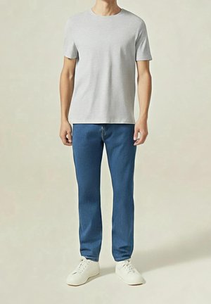 Mann trägt ein schlichtes hellgraues Kurzarm-T-Shirt, blaue Jeans und weiße Turnschuhe und steht vor einem neutralen Hintergrund.