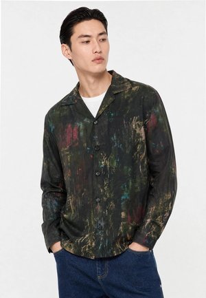 Homme portant une chemise boutonnée multicolore foncée sur un sous-vêtement blanc avec un jean bleu, debout les mains dans les poches, regardant sur le côté.