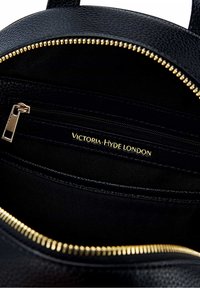 Intérieur de sac à main en cuir noir avec quincaillerie et fermetures éclair dorées, présentant une étiquette de marque "Victoria Hyde London" et une finition lisse et texturée.