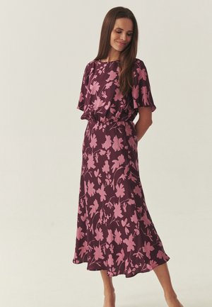 Žena stojaca so skrytými rukami za chrbtom, oblečená v fialových šatách s ružovými kvetinovými vzormi a krátkymi rukávmi na svetlom pozadí.