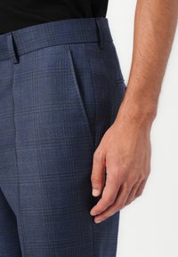 Pantalones a cuadros azul marino con una textura suave, que presentan un frente plano, trabillas para cinturón y bolsillos laterales. Mano mostrada cerca de la cintura.