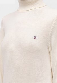 Pull à col roulé tricoté de couleur crème avec un petit logo brodé en forme de bouclier arborant des étoiles et des rayures sur la poitrine gauche.