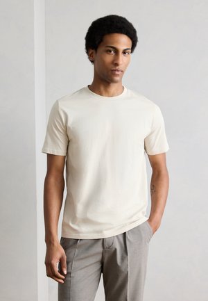 Tânăr cu păr scurt și creț, purtând un tricou simplu bej cu mâneci scurte și pantaloni gri, stând cu o mână în buzunar.