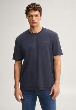 Jeune homme aux cheveux bouclés portant un t-shirt bleu foncé uni et un jean bleu, debout devant un fond neutre.