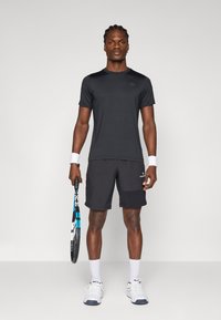 Μαύρο αθλητικό μπλουζάκι και μαύρη κοντή παντελόνι με τσέπη, κρατώντας μια ρακέτα τένις, ολοκληρωμένο με λευκές επιδερμίδες χεριών και λευκά αθλητικά παπούτσια.