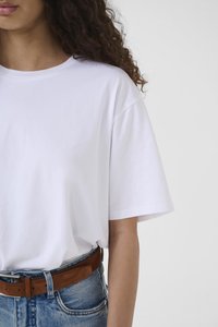 Kaaslast juustega naine, kes kannab valget lihtsat T-särki, mis on sisse püksitud sinistesse teksadesse koos pruuni seemisnahast vööga, taustal hele ühevärviline taust.