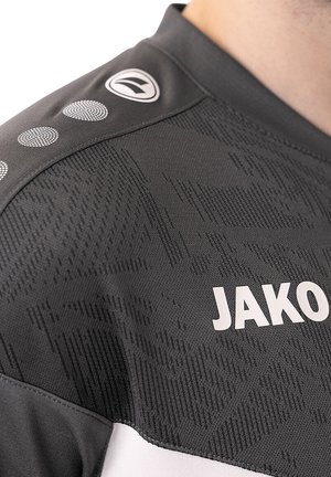 Grijs sportshirt met een textuurpatroon, witte accenten en cirkelvormige details op de schouder. Merknaam 'JAKO' in vetgedrukte witte letters.