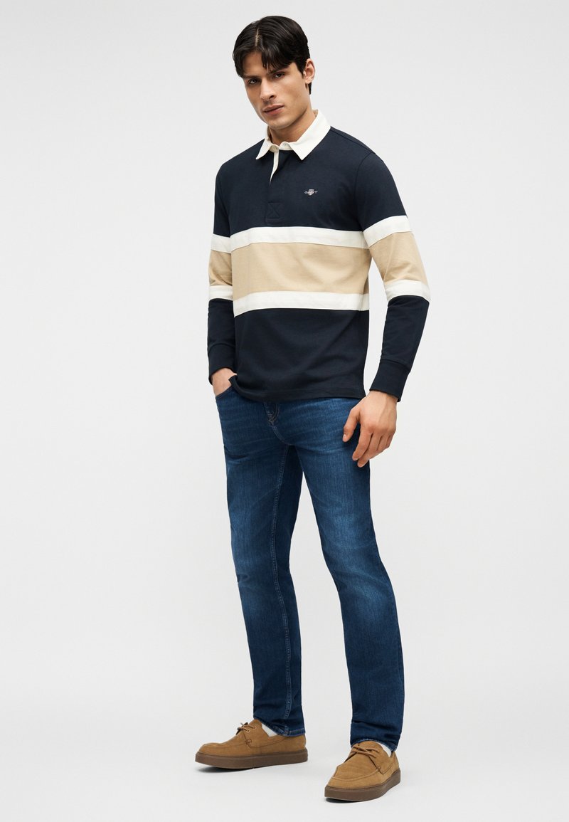 Jeune homme portant un polo à manches longues rayé bleu marine, beige et blanc, un jean bleu et des chaussures décontractées marron, debout les mains dans les poches.