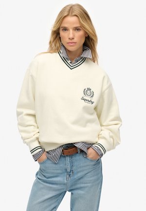 Sudadera crema con detalles en verde, con un logo, llevada sobre una camisa de cuello a rayas. Combinada con jeans de color azul claro y un cinturón marrón.