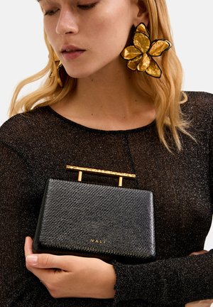 Borsa nera texturizzata con manico in metallo dorato, tenuta da un modello che indossa un top scintillante nero. La borsa mostra il nome del marchio "NALI."