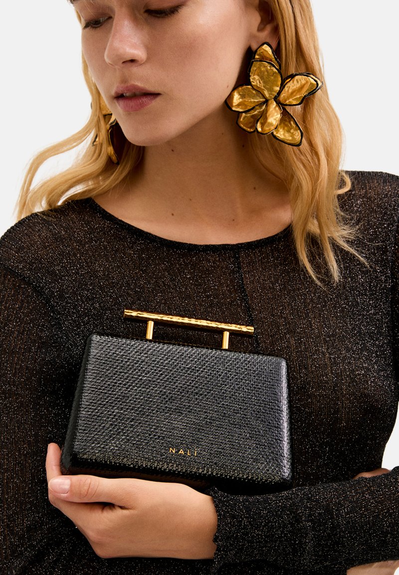 Borsa nera texturizzata con manico in metallo dorato, tenuta da un modello che indossa un top scintillante nero. La borsa mostra il nome del marchio "NALI."
