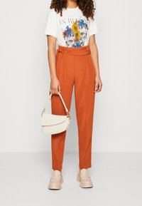 Bílé grafické tričko s květinovým potiskem spárované s oranžovými kalhotami v pase. Outfit doplňuje béžová kabelka a robustní béžové boty.