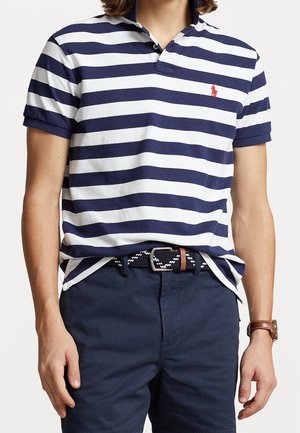 Personne portant un polo à manches courtes rayé bleu marine et blanc avec un logo rouge, un short bleu marine, une ceinture tressée et une montre marron.