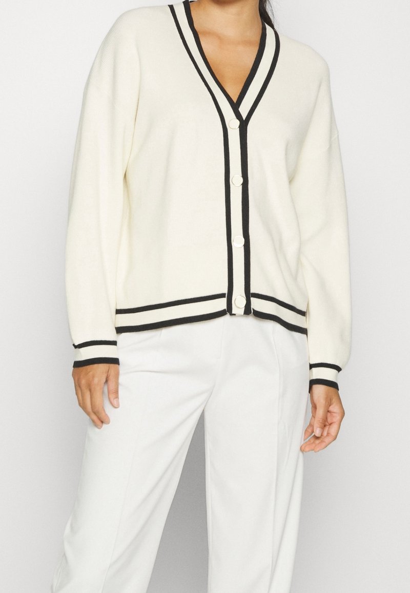 Krémový kardigan s výstřihem do V, černým lemem a pěti knoflíky. Má dlouhé rukávy a volný střih. Pairovaný s bílými kalhotami.