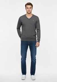 Maglione grigio scuro a scollo a V con un motivo testurizzato, indossato con jeans blu e sneakers bianche, con un piccolo logo sul petto.