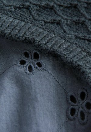 Pull en maille sombre avec un motif texturé, révélant un tissu sombre lisse en dessous avec des broderies florales et de petits détails découpés.